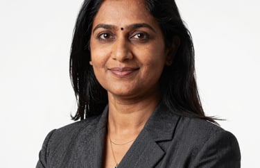 A professional studio portrait of a South Asian female educator in professional attire. She has a warm, intelligent expression. Clean, high-key lighting against a white background.