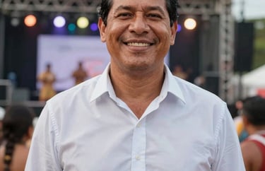 A professional South American / Brazilian Amazonian outdoor portrait cropped into a square shape of a male cultural producer in Santa Bárbara do Pará. Smiling warmly, wearing a crisp light shirt near a festive stage.