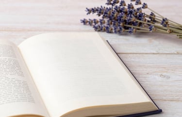 A detail of an open book on a pale wood table next to a sprig of dried lavender, soft pearl pink background, Southern European / Italian office.