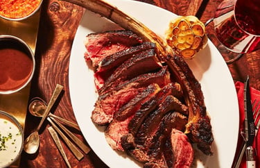 Sliced medium-rare tomahawk ribeye steak served with roasted garlic, sauces, and red wine.