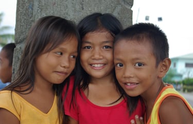 Three smiling Filipino children posing closely together outdoors in a rural village setting.