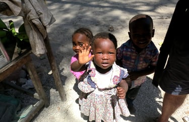 Three young African children standing outside and smiling, with one girl waving her hand.