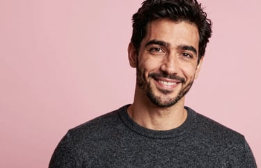 A headshot of an International / English-speaking professional man with a friendly expression, wearing a dark grey sweater, standing against a soft light pink background.
