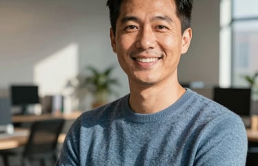 A professional headshot of a person in a muted blue sweater, smiling confidently in a sunlit modern workspace. North American / US urban office setting.