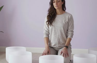 A professional portrait of a sound healer standing next to large crystal bowls in a North American holistic center. The room is decorated in soft white and pale lavender tones.