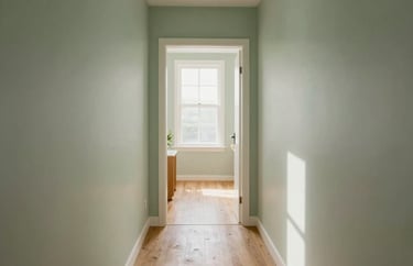 A bright hallway with light wood floors and soft sage green walls, leading toward a sun-drenched common area, photography.