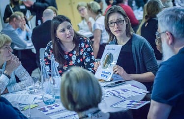 A woman in glasses holding a Fiona's Story pamphlet during a mental health workshop table discussion.