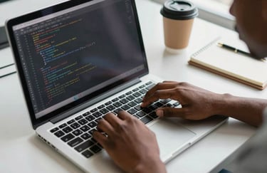Close-up overhead shot of a young African developer's hands on a laptop keyboard in a bright tech lab, lines of code visible on screen, natural daylight from a side window, a coffee cup and notebook nearby