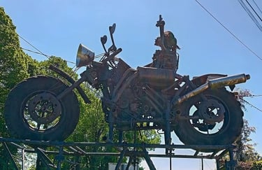 A large rusty metal motorcycle sculpture made from scrap parts displayed outdoors against a blue sky.