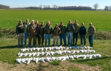 Ten hunters in various outdoor gear posing behind four long rows of harvested snow geese in a green Maryland field.