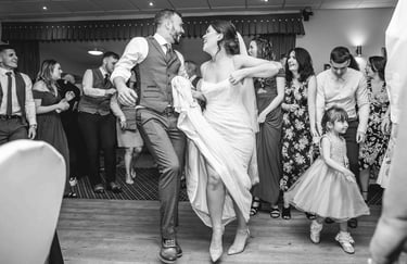 Black and white photo of a happy bride and groom dancing with wedding guests at a reception.