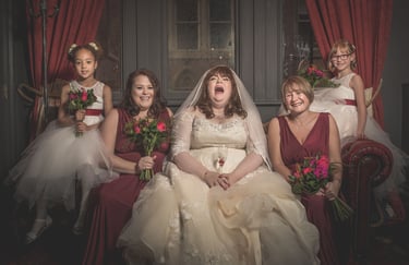 A laughing bride in a lace gown sits with bridesmaids in burgundy dresses and flower girls holding bouquets.