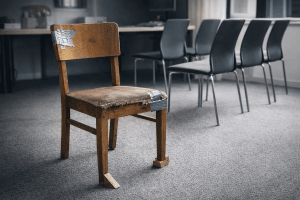 A repaired, unstable wooden chair used despite modern office chairs sitting unused in the background