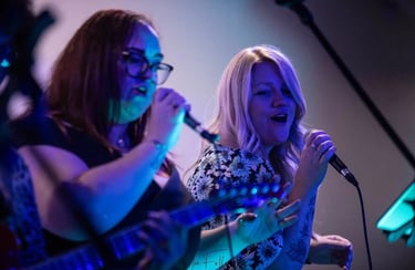 Two female vocalists performing live music on stage with microphones and blue stage lighting.