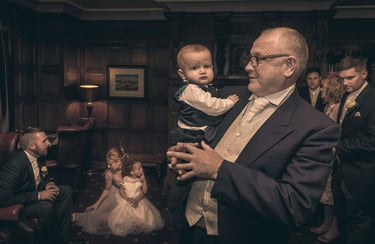 Grandfather in a formal suit holding a baby boy at a wedding reception in a wood-paneled room.