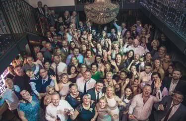 A large group of wedding guests smiling and waving at a party under a crystal chandelier.