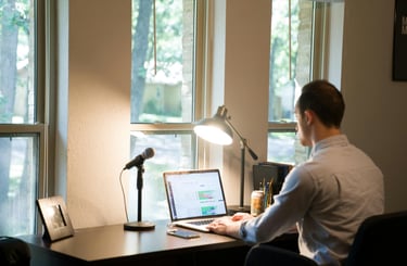 A man working at a home office desk with a laptop, microphone, and lamp by a window.