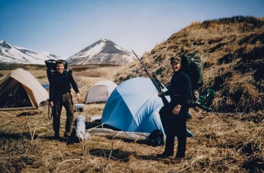 Two hunters at a remote Alaskan base camp with tents and mountains in the background.