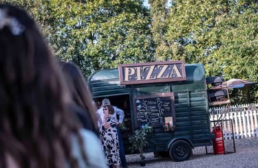 Rustic horsebox pizza trailer serving guests at an outdoor wedding event with a chalkboard menu.