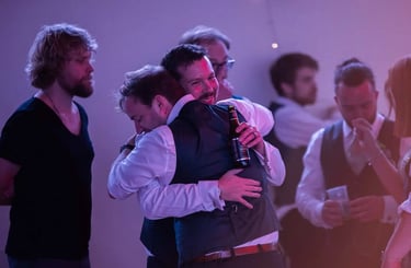 Smiling groom hugging a friend at a wedding reception party with guests and drinks in the background.