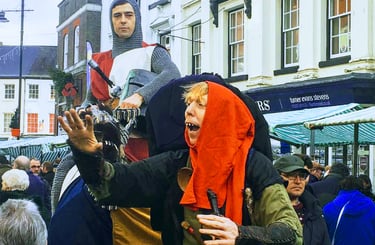 Performers in medieval costumes, including a knight and jester, entertain a crowd at an outdoor festival.