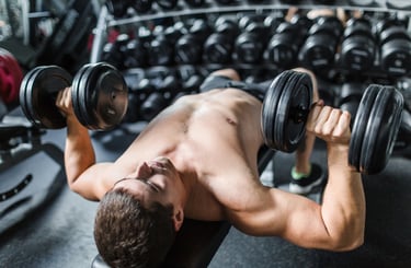 A shirtless man performing a dumbbell chest press on a weight bench in a gym setting.