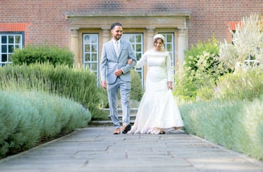 asian bride & groom outside country house walking towards camera linking arms and smiling, bushes are leading lines