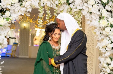 candid photo of Somalian groom kissing bride on forehead, his hugging her tight, floral arch framing them