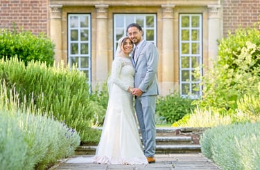asian bride and groom holding hands and looking at camera I'm front of a Stately home, bushes are leading lines 