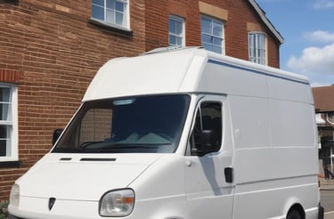 A FedEx Express delivery van is parked on the side of a street. The foreground features a road with yellow and white lane markings. In the background, there are several tall leafy trees and a row of commercial buildings, some with red brick facades. A small park or public square is visible, with people walking and a few tents or stalls set up.