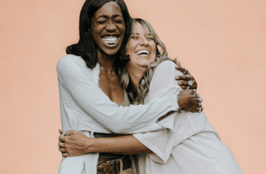 Twee vrouwen lachen en omhelzen elkaar hartelijk tegen een zachte perzikkleurige achtergrond. Ze stralen warmte, verbondenhei