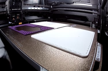 A Bambu Lab 3D printer creating purple and white plastic parts on a heated build plate.