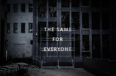 Illuminated neon sign saying The Same For Everyone on metal scaffolding in front of a modern office building.
