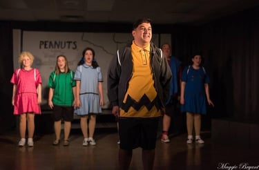 an actor playing Charlie Brown in a yellow sweater and black shorts standing in front of a group of