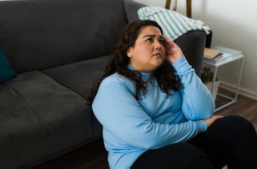 A stressed woman sitting on the floor leaning against a gray sofa, touching her head due to a headache.