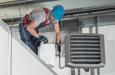 A HVAC technician in safety gear installs a commercial ceiling mounted air heater.