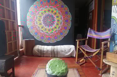 Sala con planta y mandala pintado en la pared, hay una silla mecedora y otros muebles rústicos con luz cálida