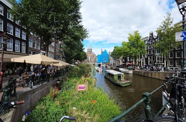 Panoramic view of an Amsterdam canal with a green tour boat, historic canal houses, and outdoor cafes.