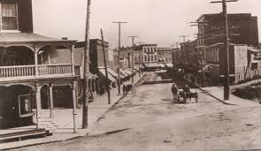 Vintage black and white photograph of a historic American main street with horse-drawn carriages and wooden buildings.