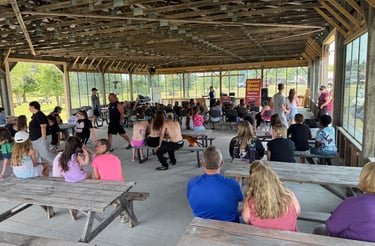 Families gather at picnic tables under a pavilion for a live outdoor magic show performance.