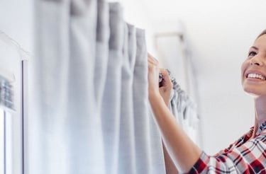 Smiling woman hanging grey blackout curtains on a window rod during home improvement.