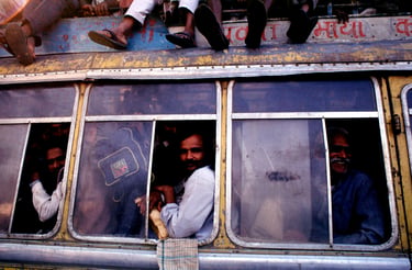 A side view of a heavily crowded yellow bus with passengers leaning out of windows and sitting on the roof.