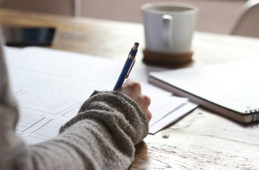A woman sits at a wooden desk, writing with pen and paper.