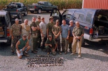 A large group of hunters in outdoor gear posing with harvested doves in a gravel lot next to several pickup trucks.