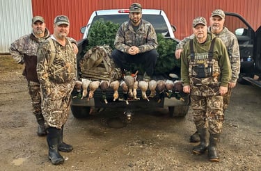 Five hunters in camouflage gear posing with harvested ducks on the tailgate of a white pickup truck.