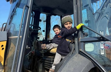 Two young boys playing inside the driver cab of a yellow John Deere construction excavator.