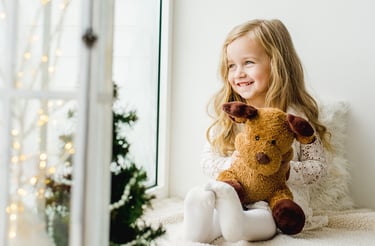 une petite fille qui regarde par la fenetre assise sur un canapé avec un ours en peluche sapin