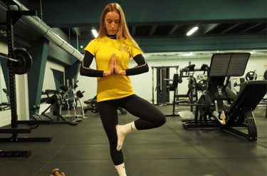Una donna in posa di yoga albero (Vrksasana) su un tappetino da yoga in palestra. Indossa una maglietta gialla con logo 'Move