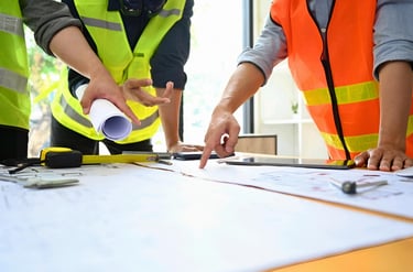 Construction engineers in safety vests reviewing architectural blueprints and site plans on a table.