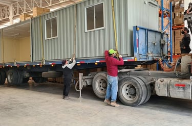 Workers securing a gray prefabricated container office onto a flatbed semi-truck for transport inside a warehouse.